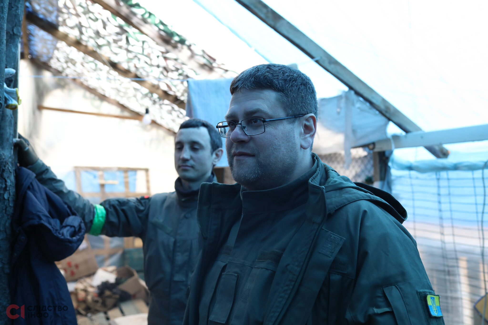 Попереду стоїть чоловік у формі й в окулярах для зору, позад нього - інший чоловік у формі з зеленою сторічкою на правій руці