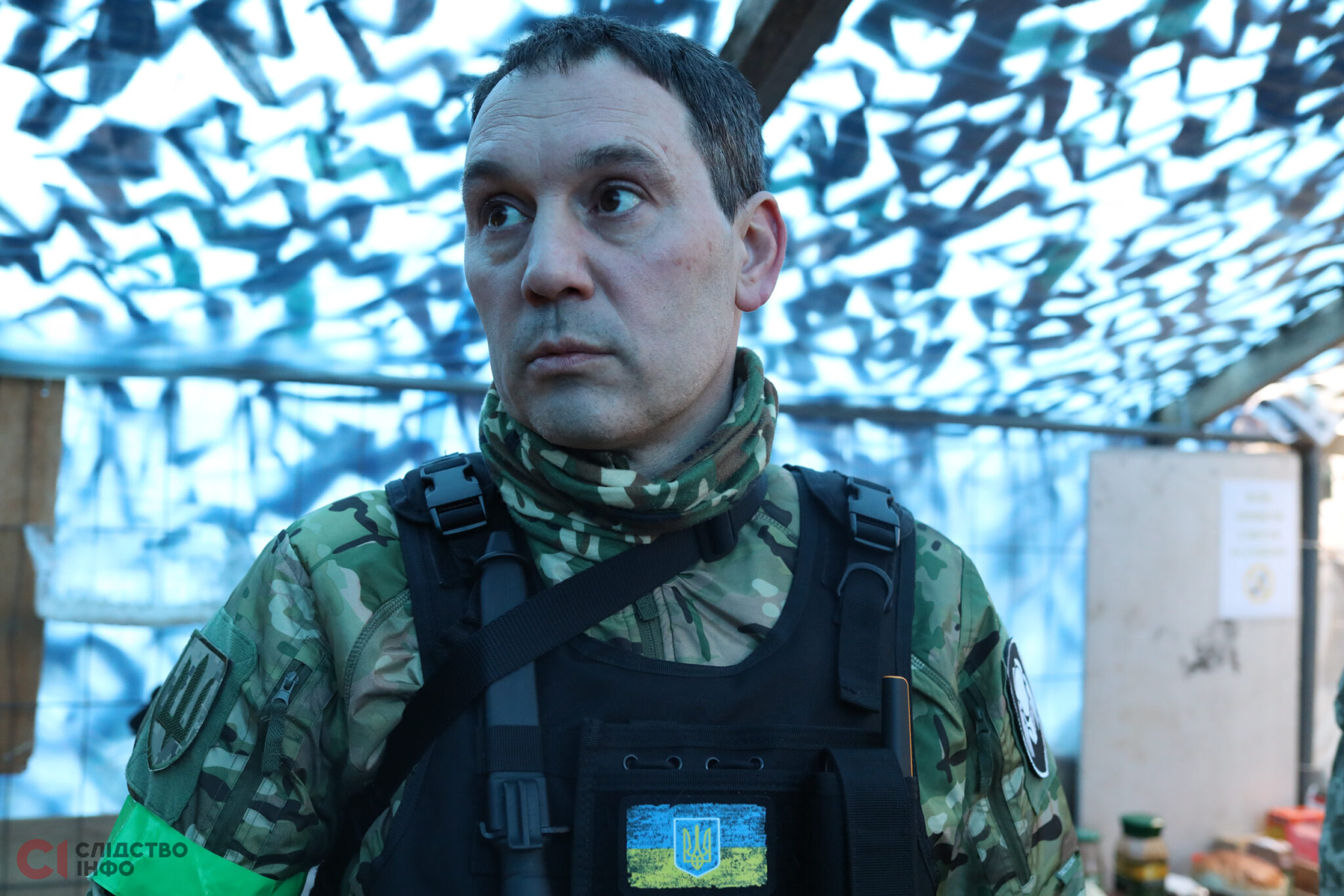 чоловік у військовій формі з шевроном України дивиться у бік, на фоні натягнута на стелі маскувальна сітка