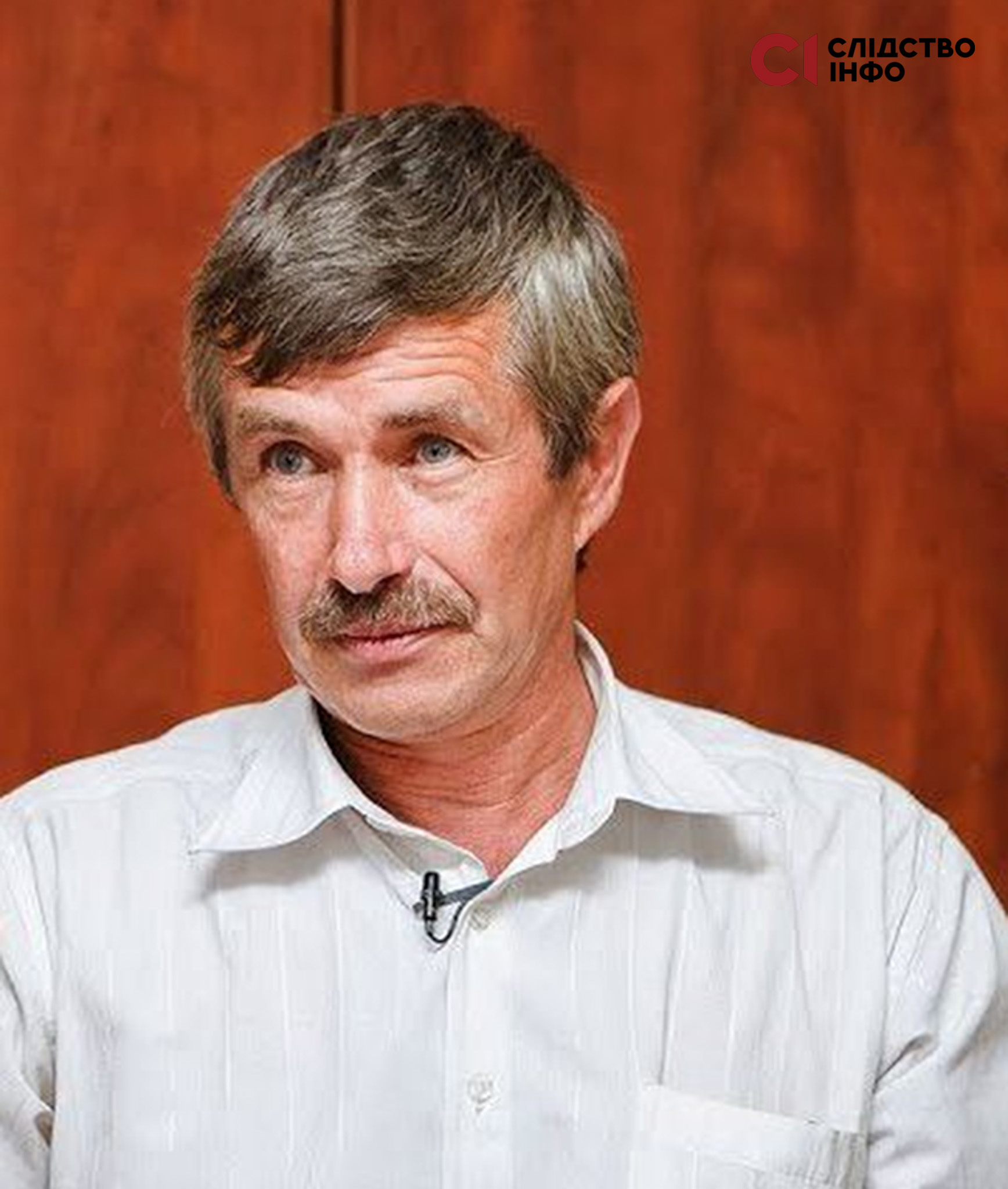 На фото вусатий чоловік у білій сорочці, позад нього багряний фон