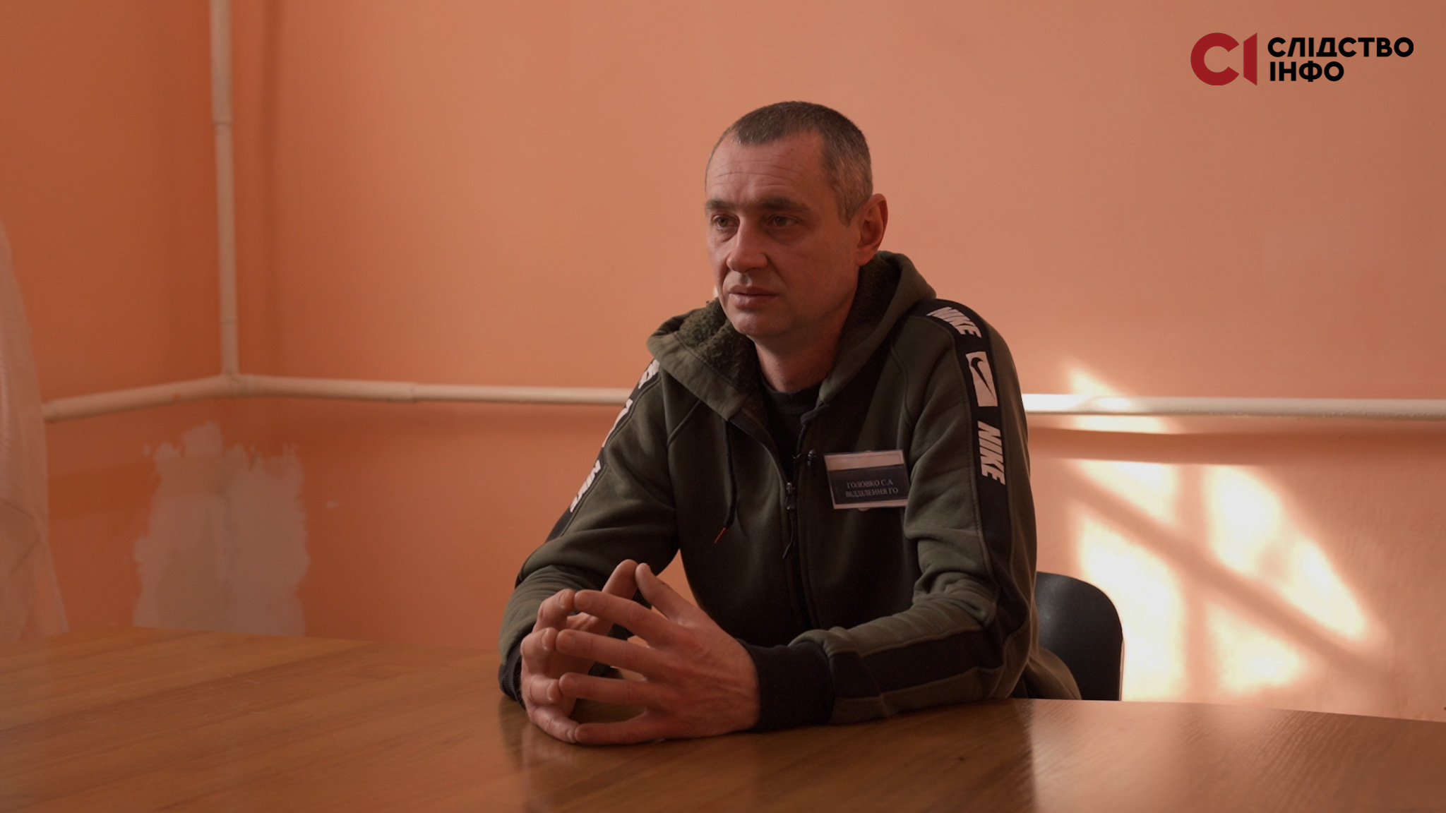 На фото чоловік у спортивній формі сидить за столом у кімнаті з рожевими стінами
