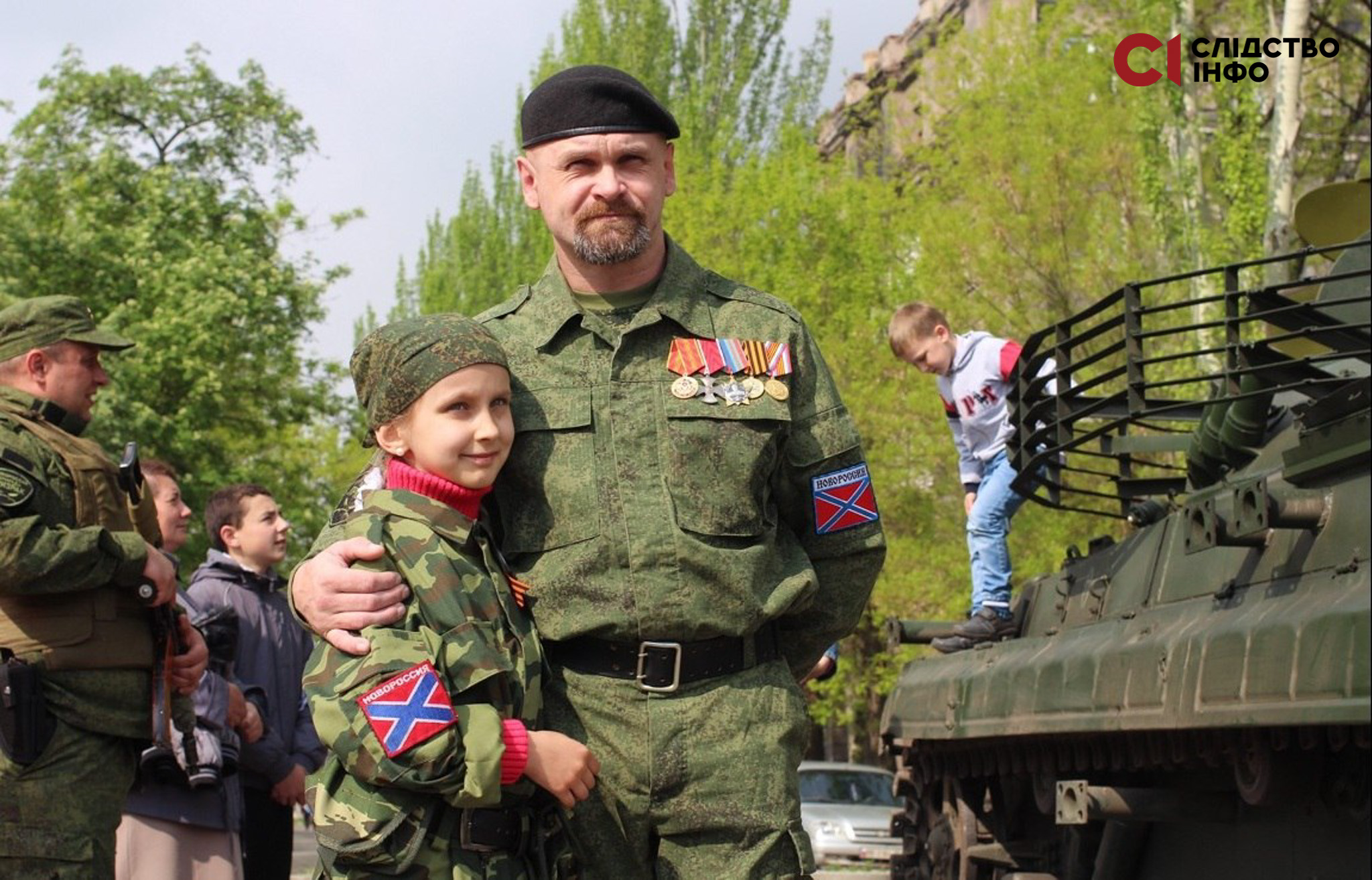 На фото чоловік у російській військовій формі обіймає маленьку дівчинку у військовій формі. Праворуч стоїть танк, на якому знаходиться маленький хлопчик. На фото є інший чоловік у