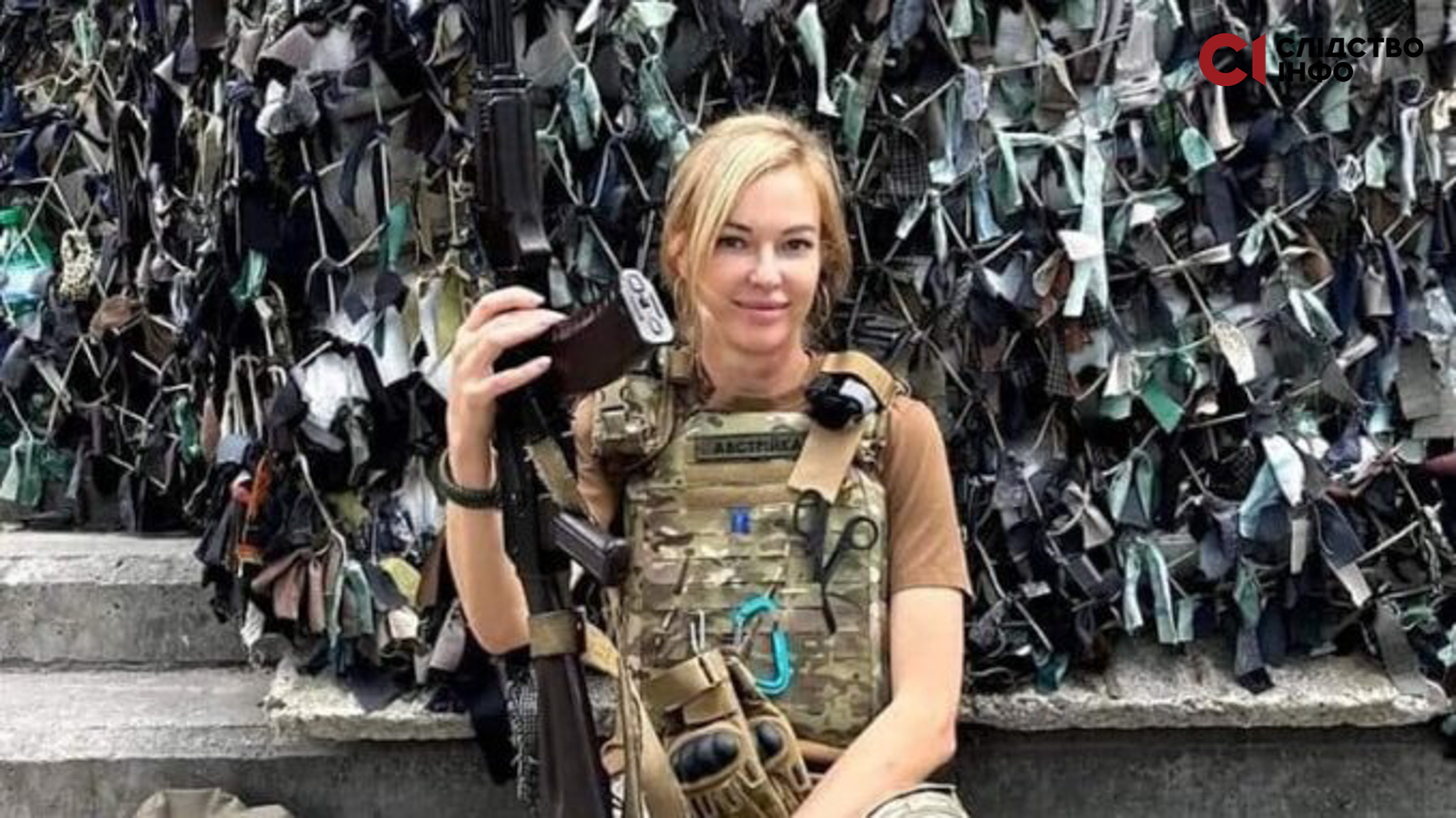 Жнка з білявим волоссям стоїть у військовій формі з піднятим догори автоматом на фоні маскувальної сітки