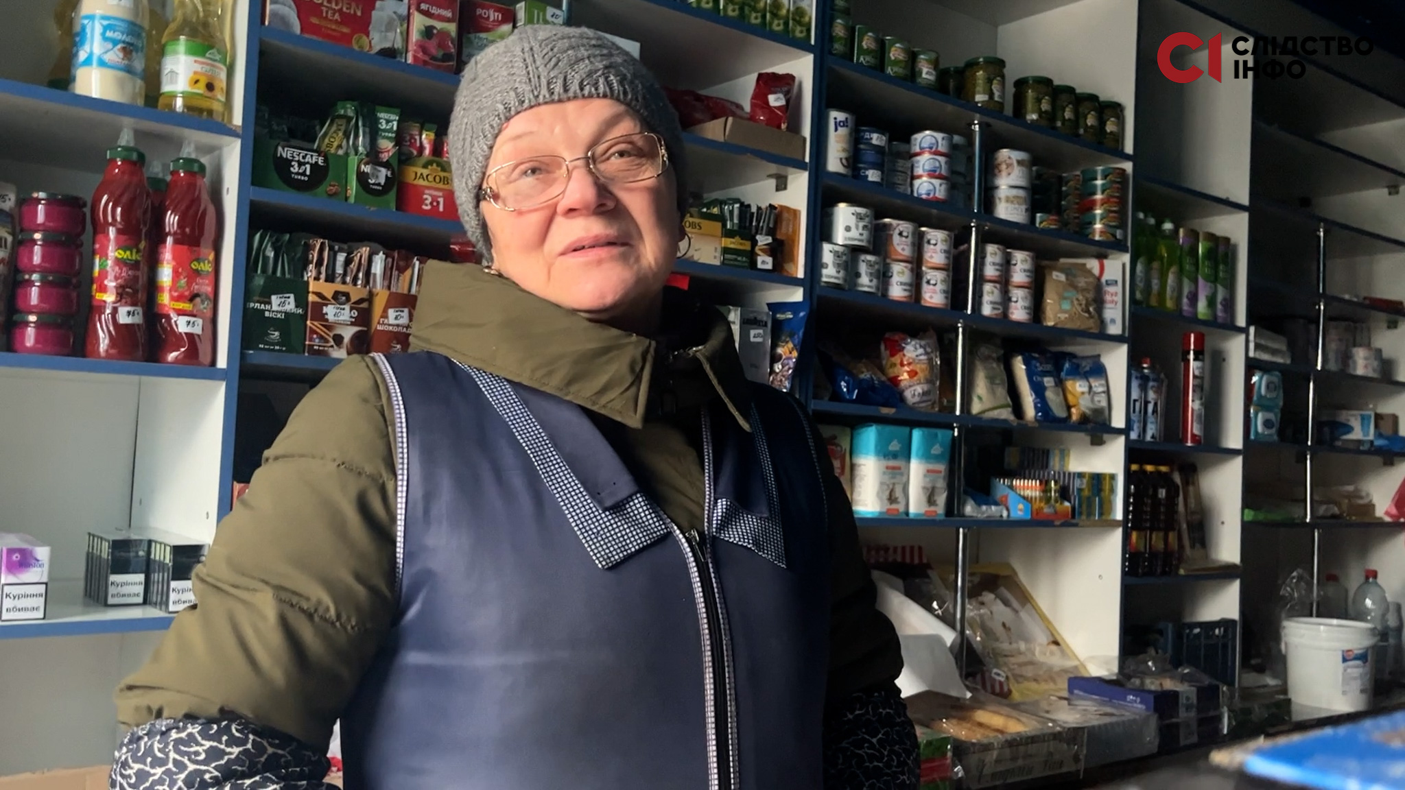 Продавщиця магазину в окулярах і шапці, позад неї полиці з продуктами