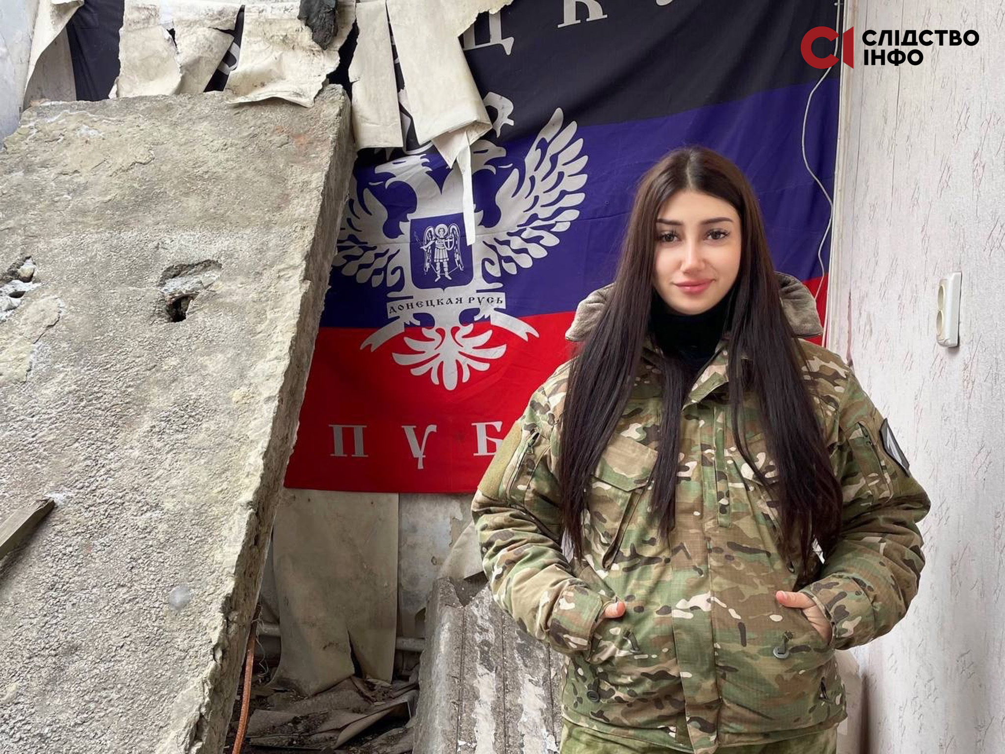 На фото 20-річна дівчина з чорним довгим волоссям у військовій формі стоїть на фоні зруйнованих стін. На стіні висить чорно-синьо-червоний правор з двоголовим білим орлом та написо