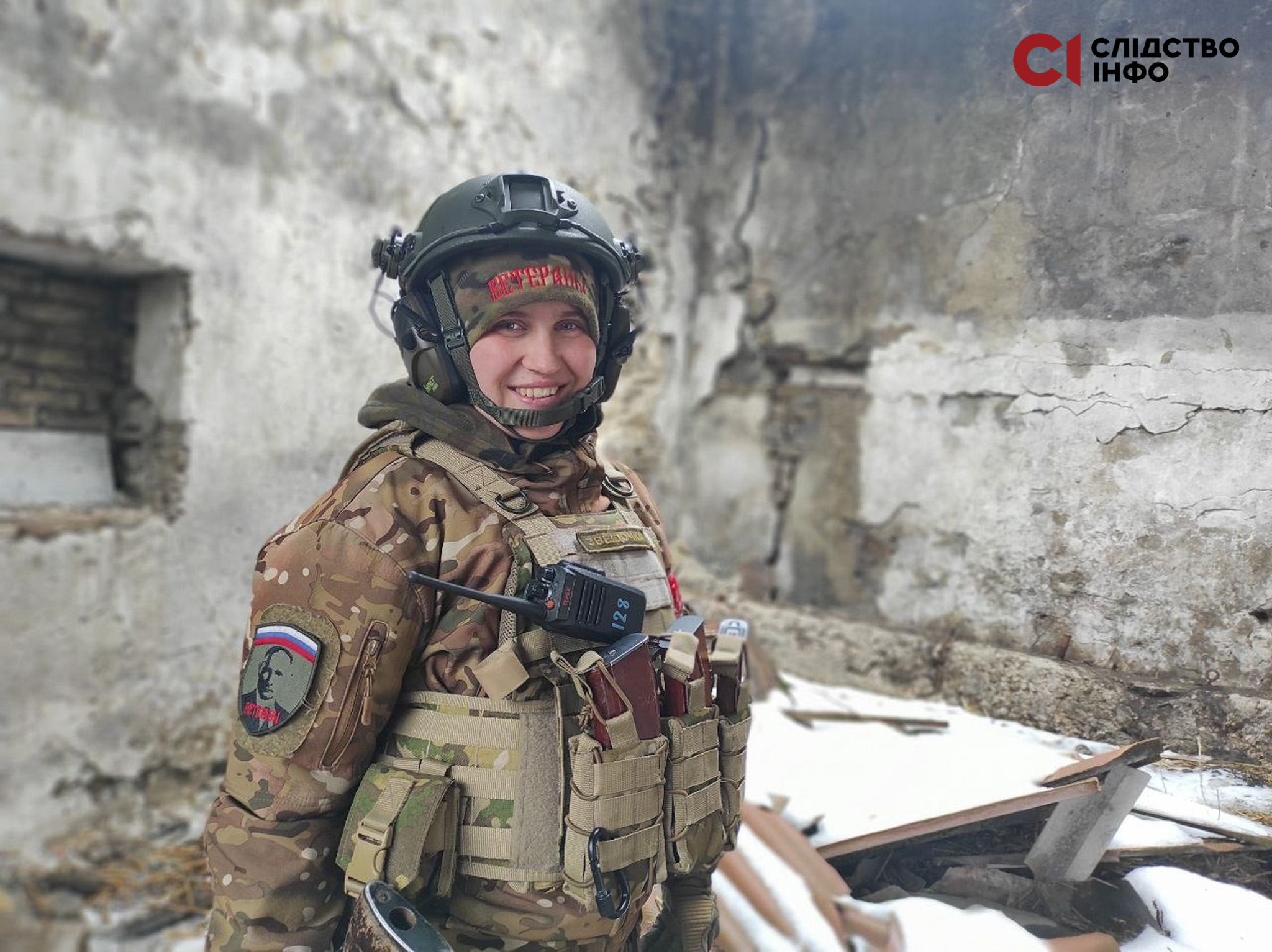 На фото молода дівчина усміхається на камеру. Вона одягнена у російську військову форму та каску, стоїть на фоні сірих стін зруйнованої будівлі. На руці у дівчини шефрон з портрето