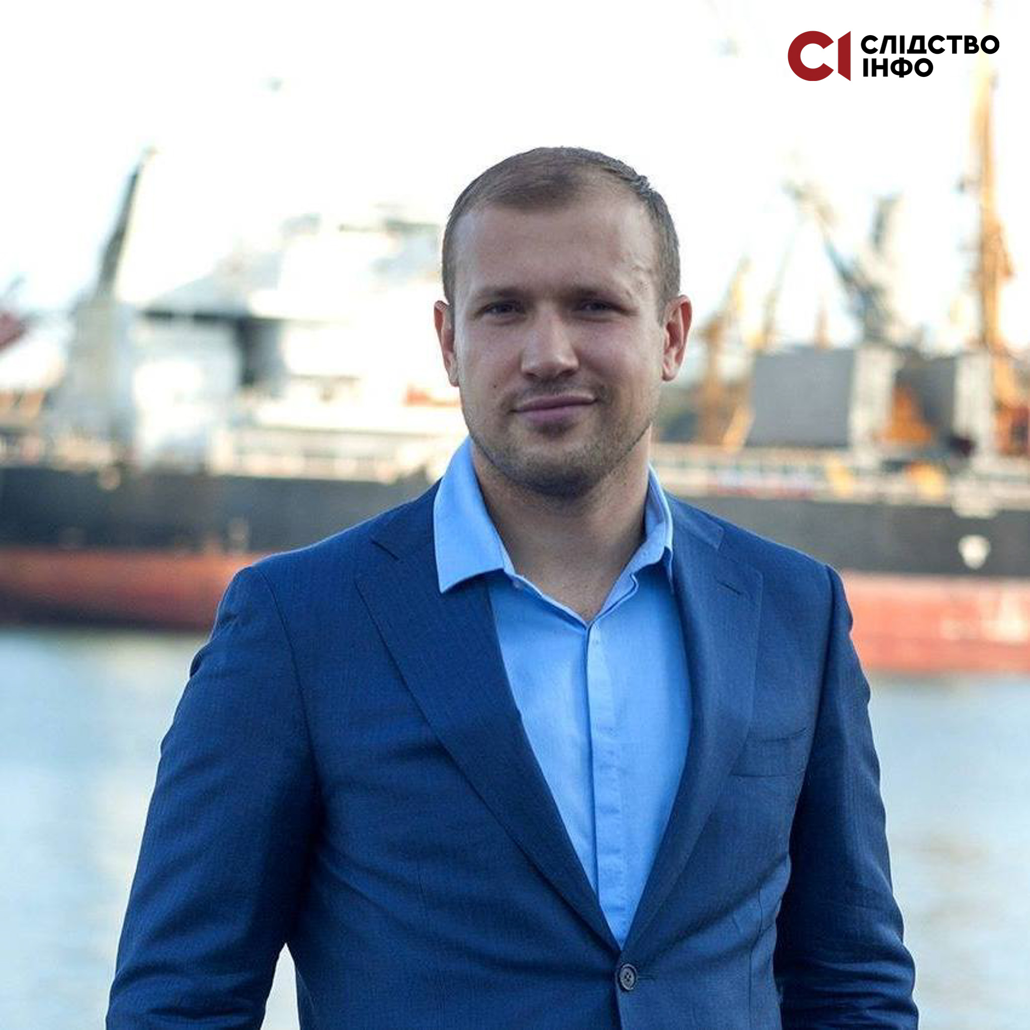 На фото чоловік у синьому костюмі й блакитній сорочці стоїть на фоні моря й кораблів