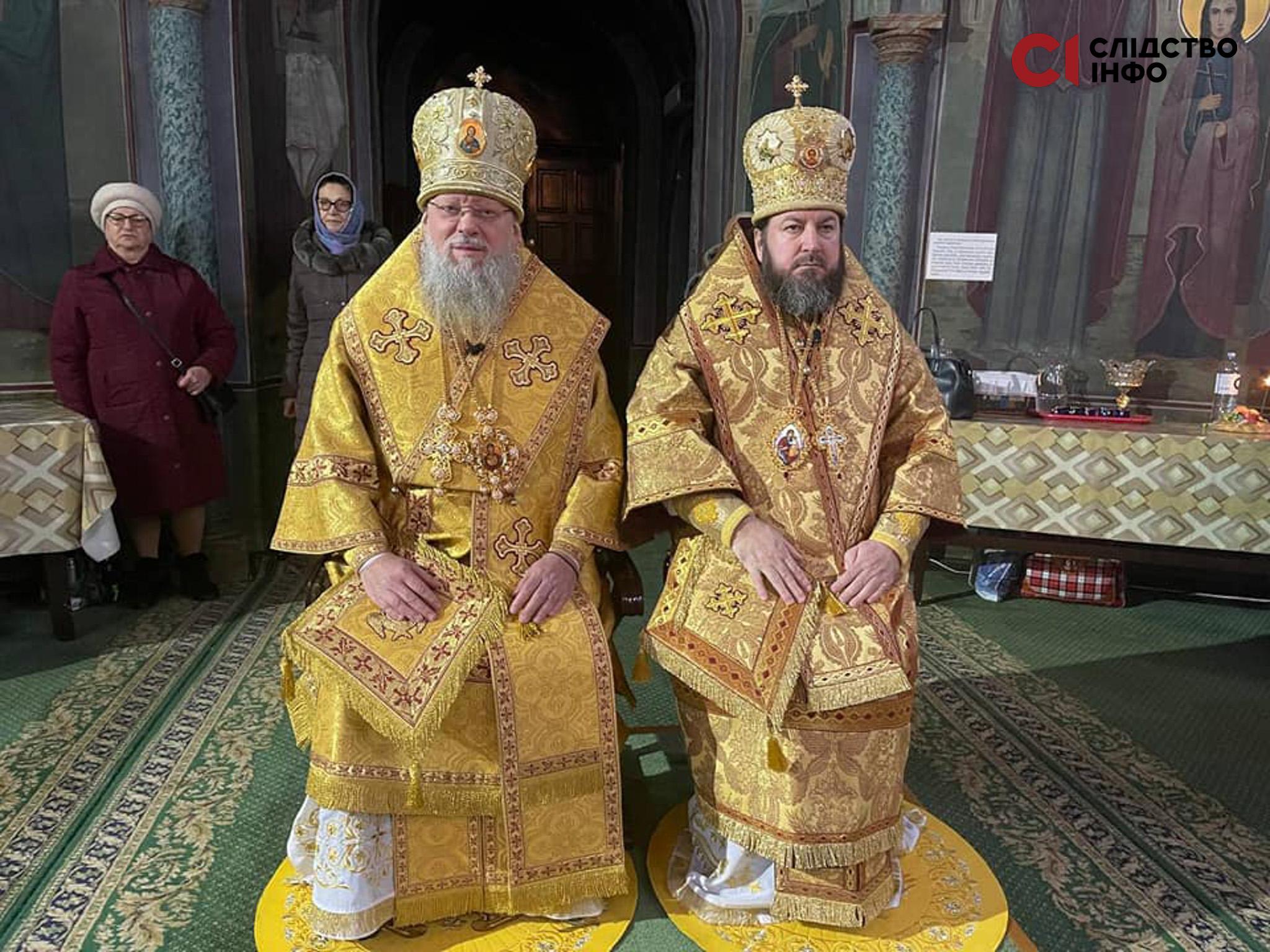 На фото сидять два священника РПЦ в Україні у золотих святкових рясах, позад них зліва стоять дві жінки в окулярах