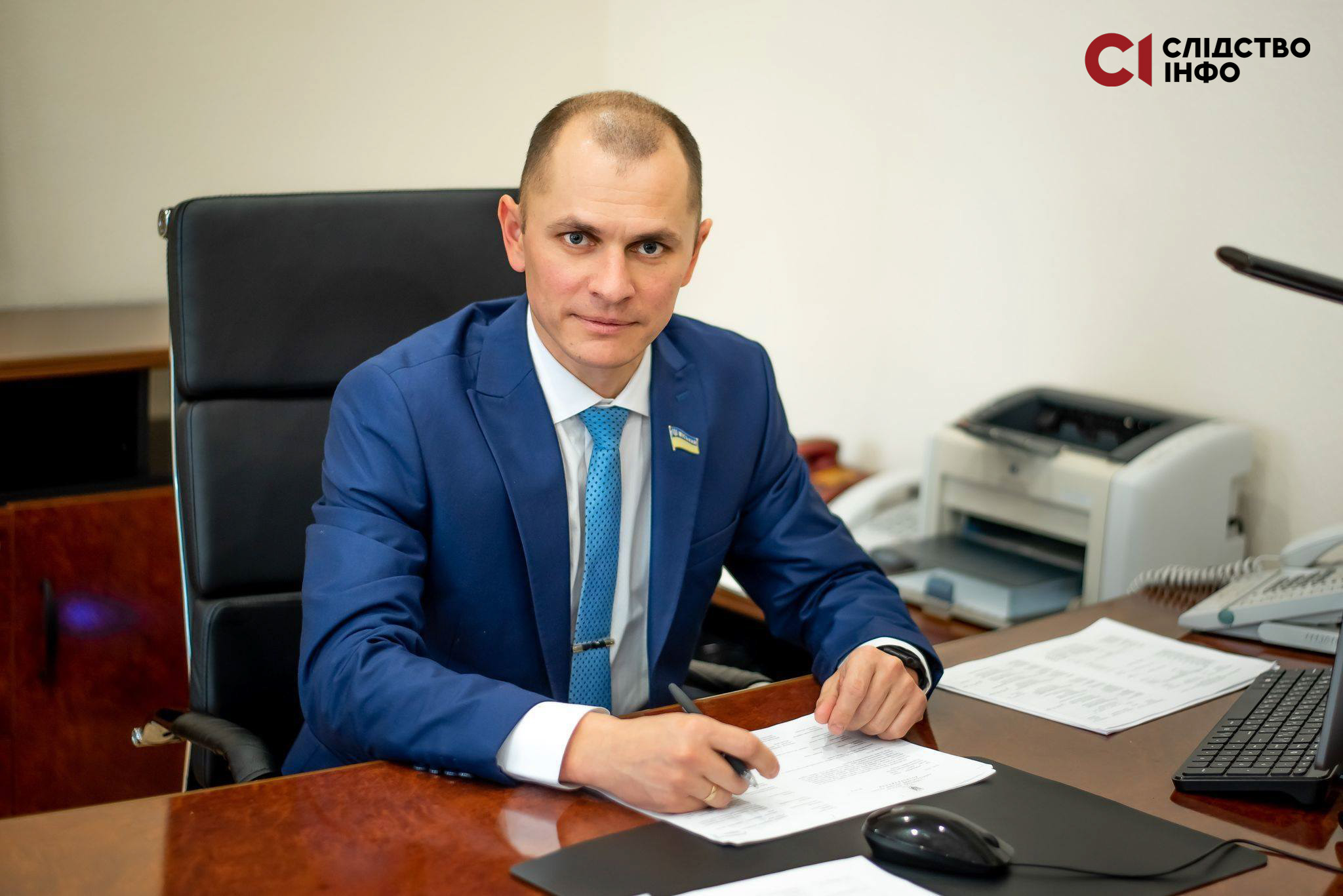 На фото чоловік у синьому костюмі сидить за столом. На столі лежать папери, праворуч стоїть принтер.