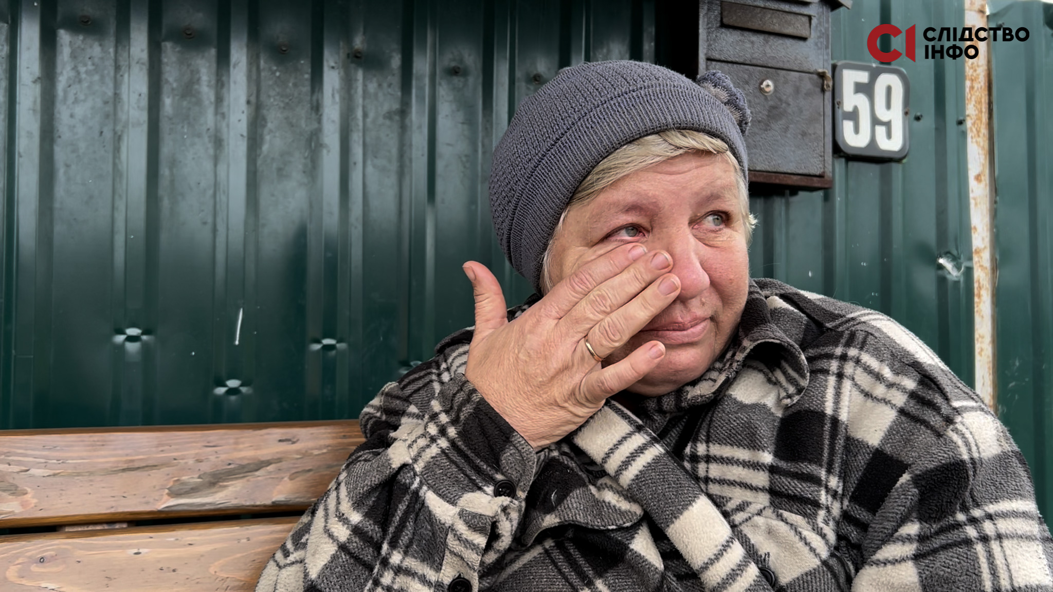 Жінка сидить на лавці й рукою витирає сльози. Позад неї темно-зелена стіна і номер будинку 59