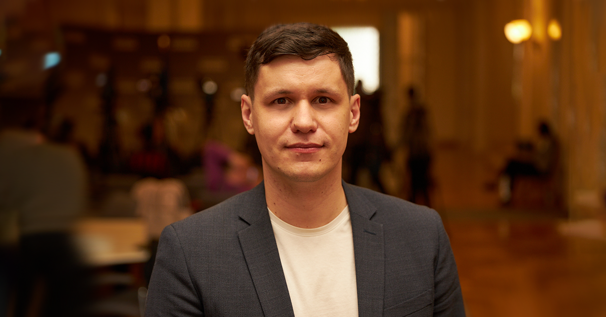 чоловік у білій футболці й сірому піджаку стоїть у приміщенні