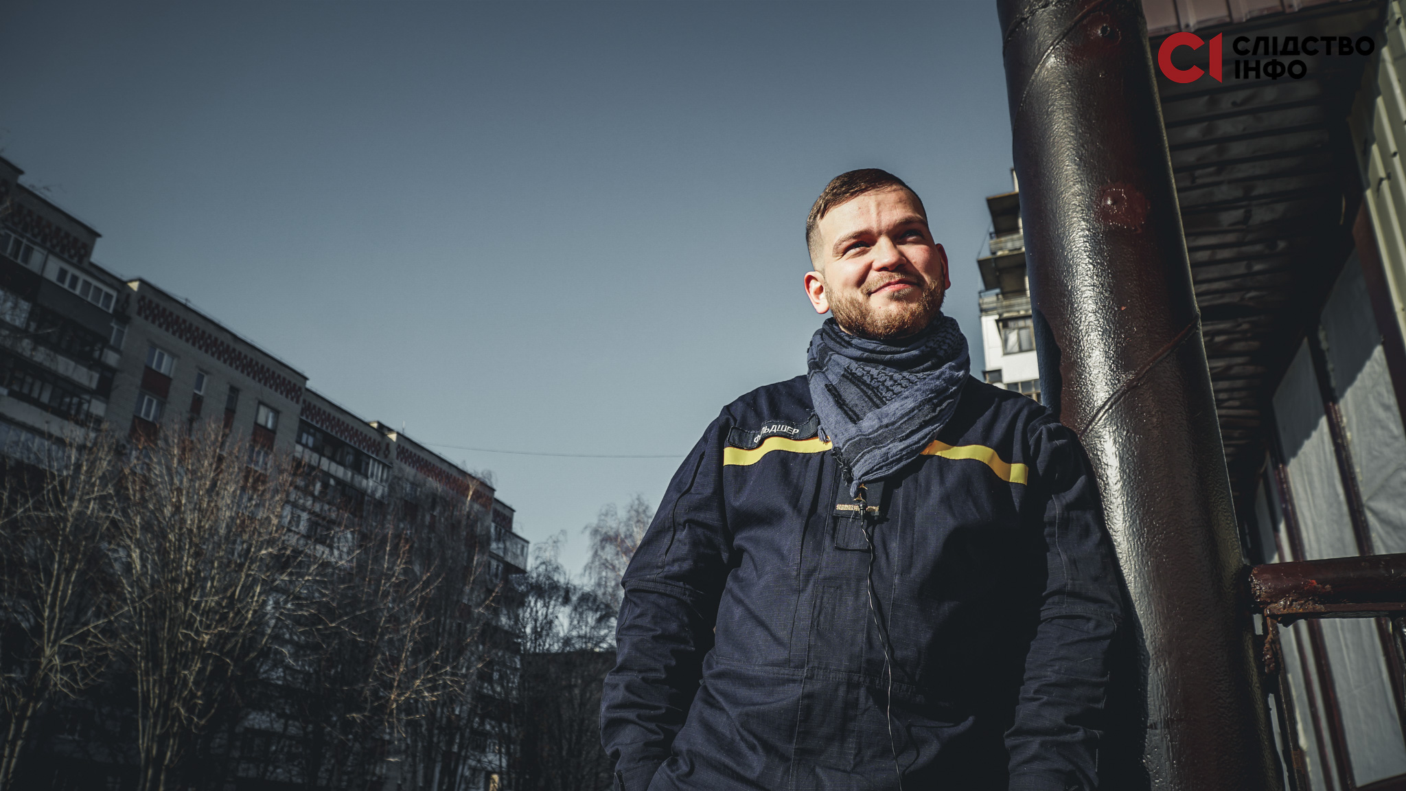 Фото усміхненого чоловіка, який притулився до великої труби, на фоні - багатоповерхівки й дерева