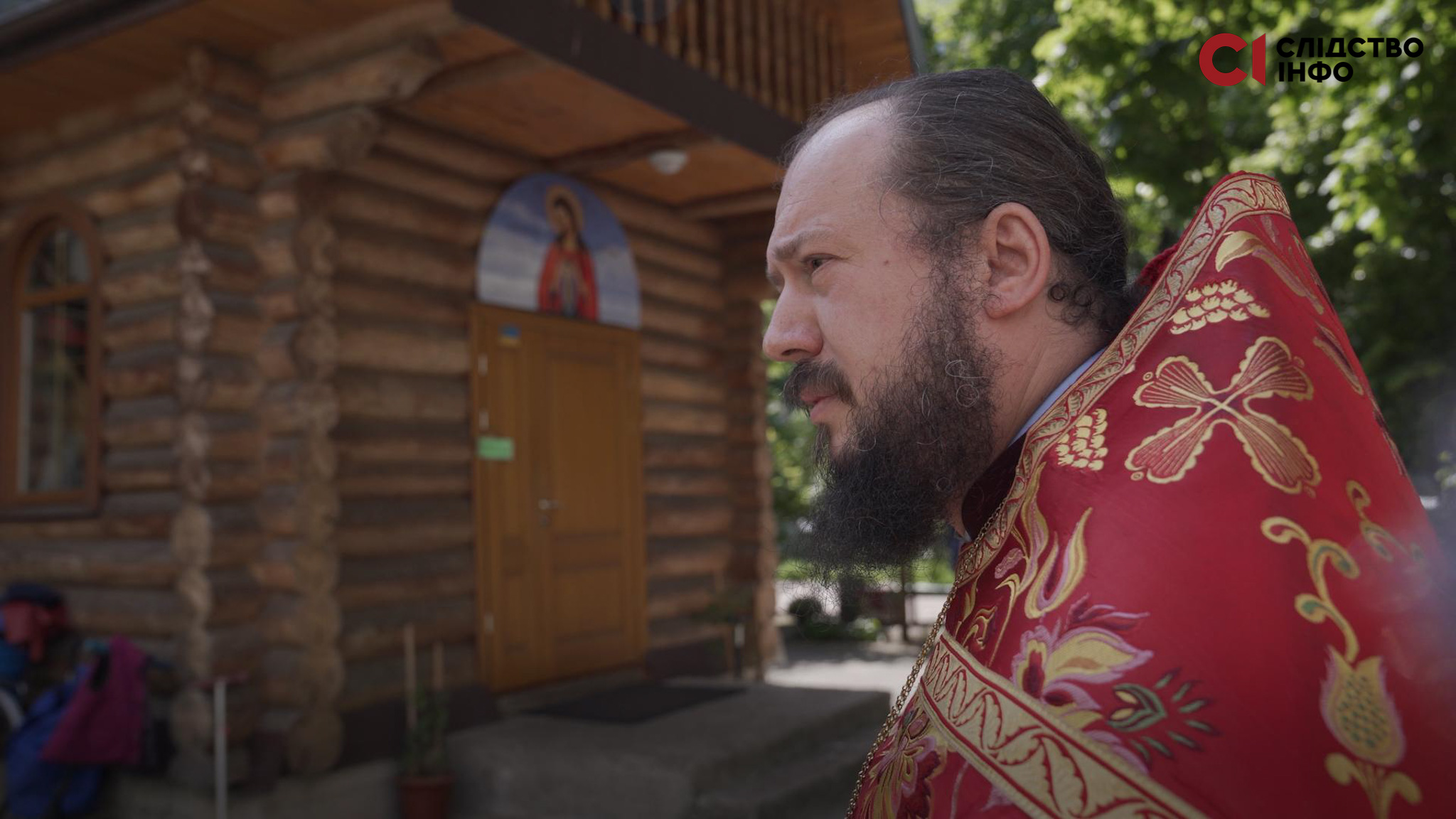 На фото священник у червоній рясі, стоїть боком на фоні дерев'яної церкви
