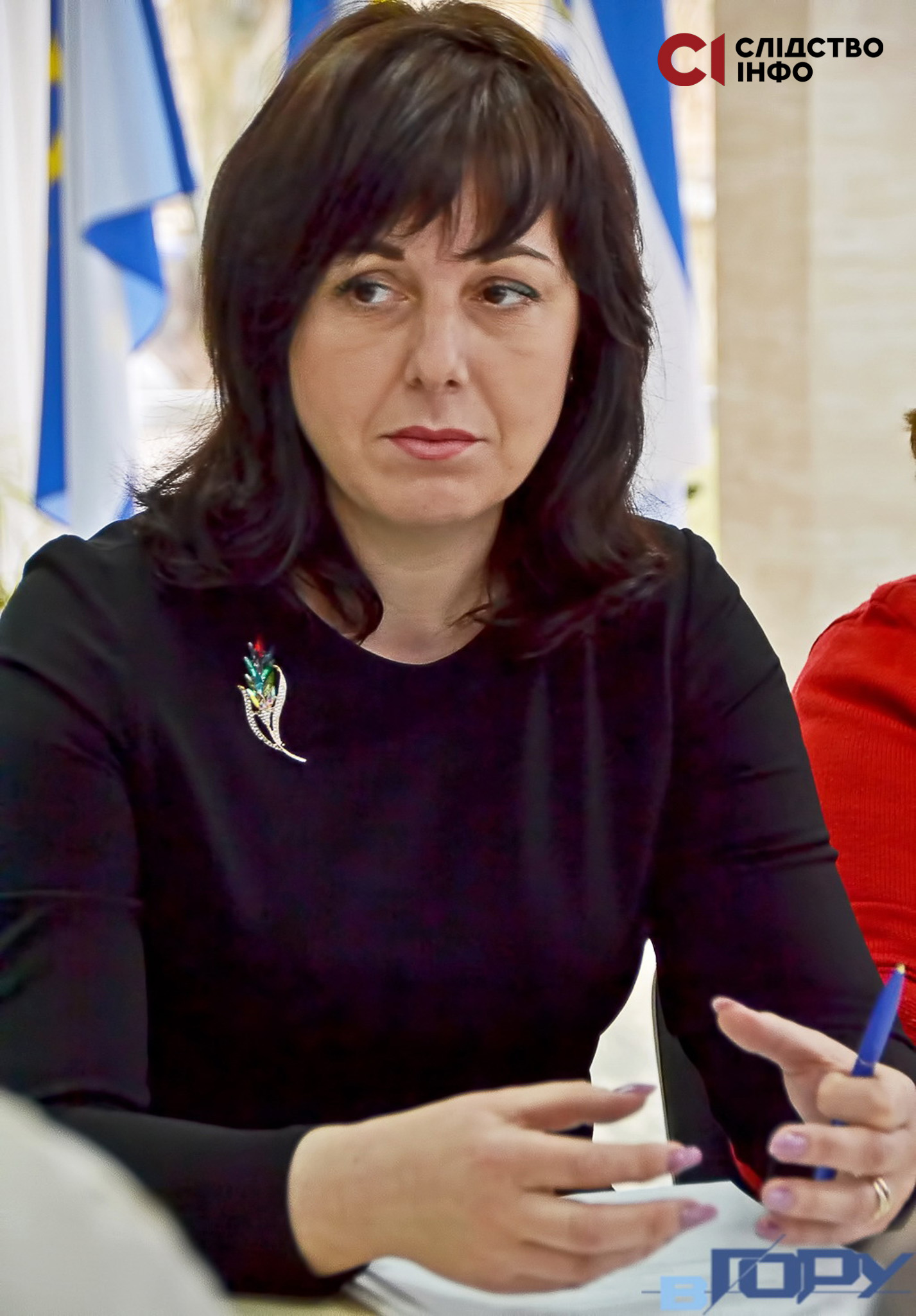 На фото жінка з темним волоссям і у чорній сукні жестикулює руками, тримаючи кулькову ручку