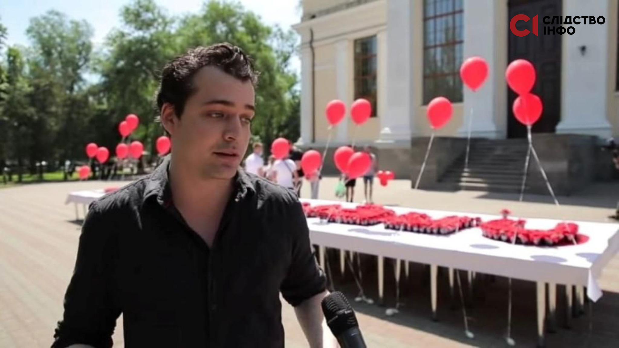 На фото чоловік у чорній сорочці стоїть на вулиці. Праворуч від нього столи з білими скатертинами. На них багато червоних кульок