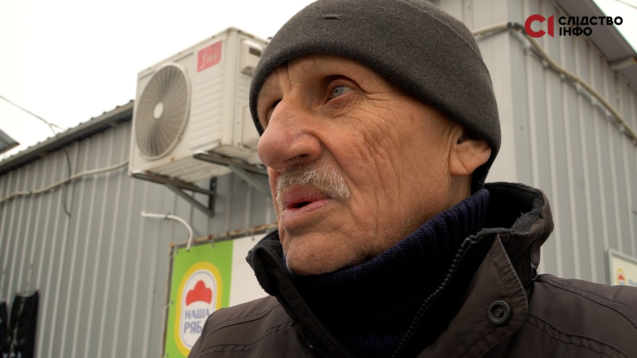 Чоловік у шапці дивиться в бік. Позад нього сіра будівля, на стіні якої прикріплений білий кондиціонер