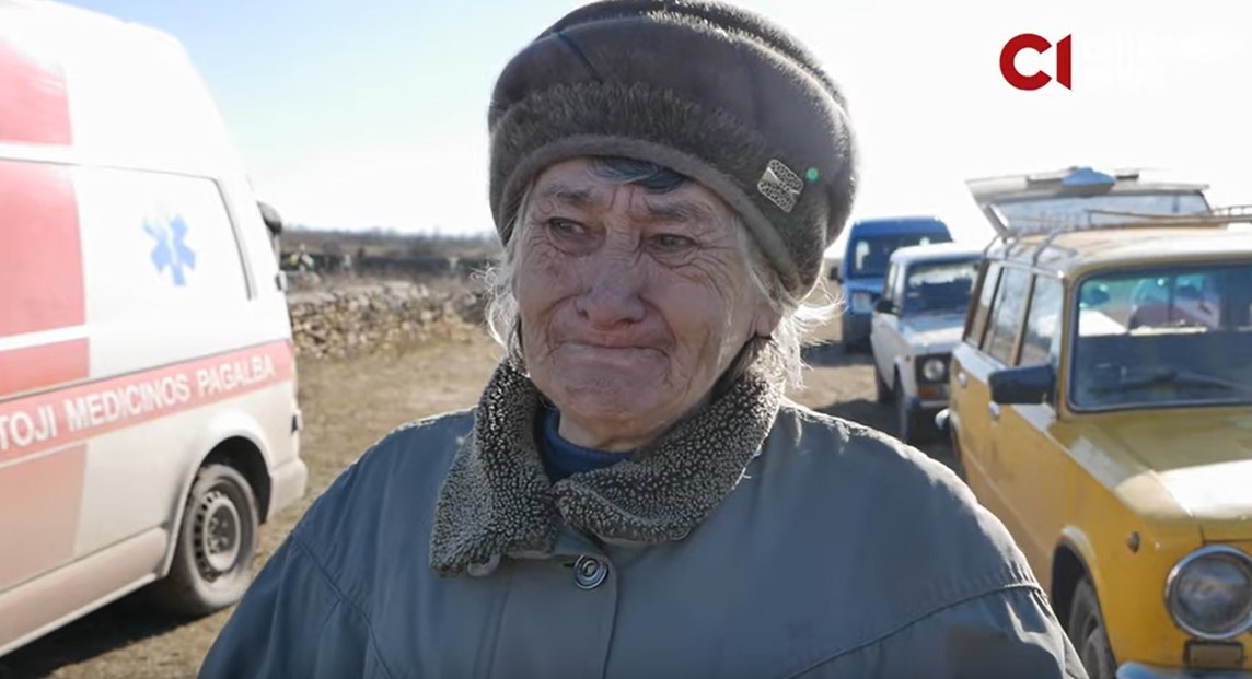 На фото старша жінка стоїть на вулиці, на фоні зліва стоїть машина швидкої допомоги, а справа - легкова машина жовтого кольору