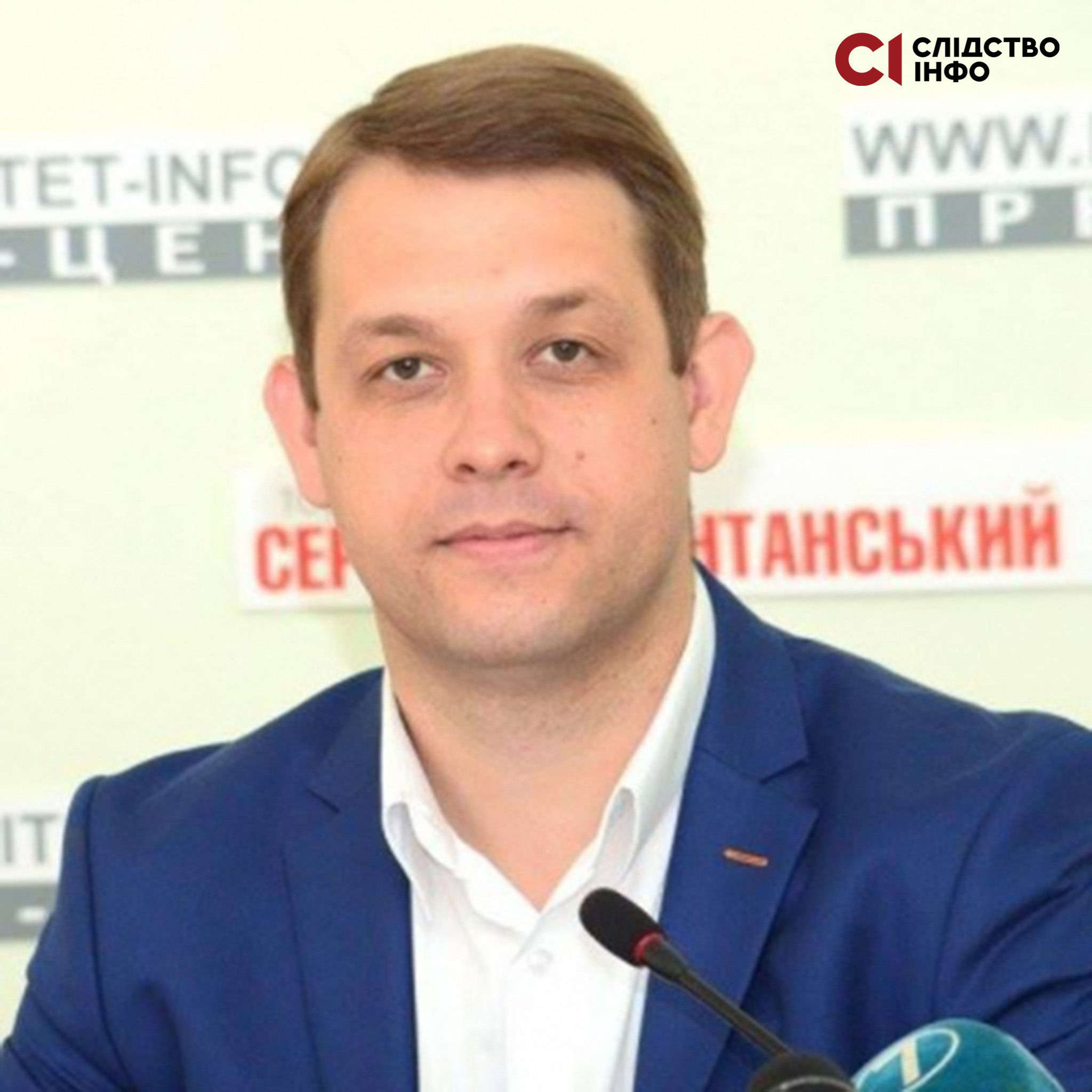 На фото чоловік у синьому піджаку й білій сорочці. Стоїть біля мікрофона на фоні світлої стіни з написами