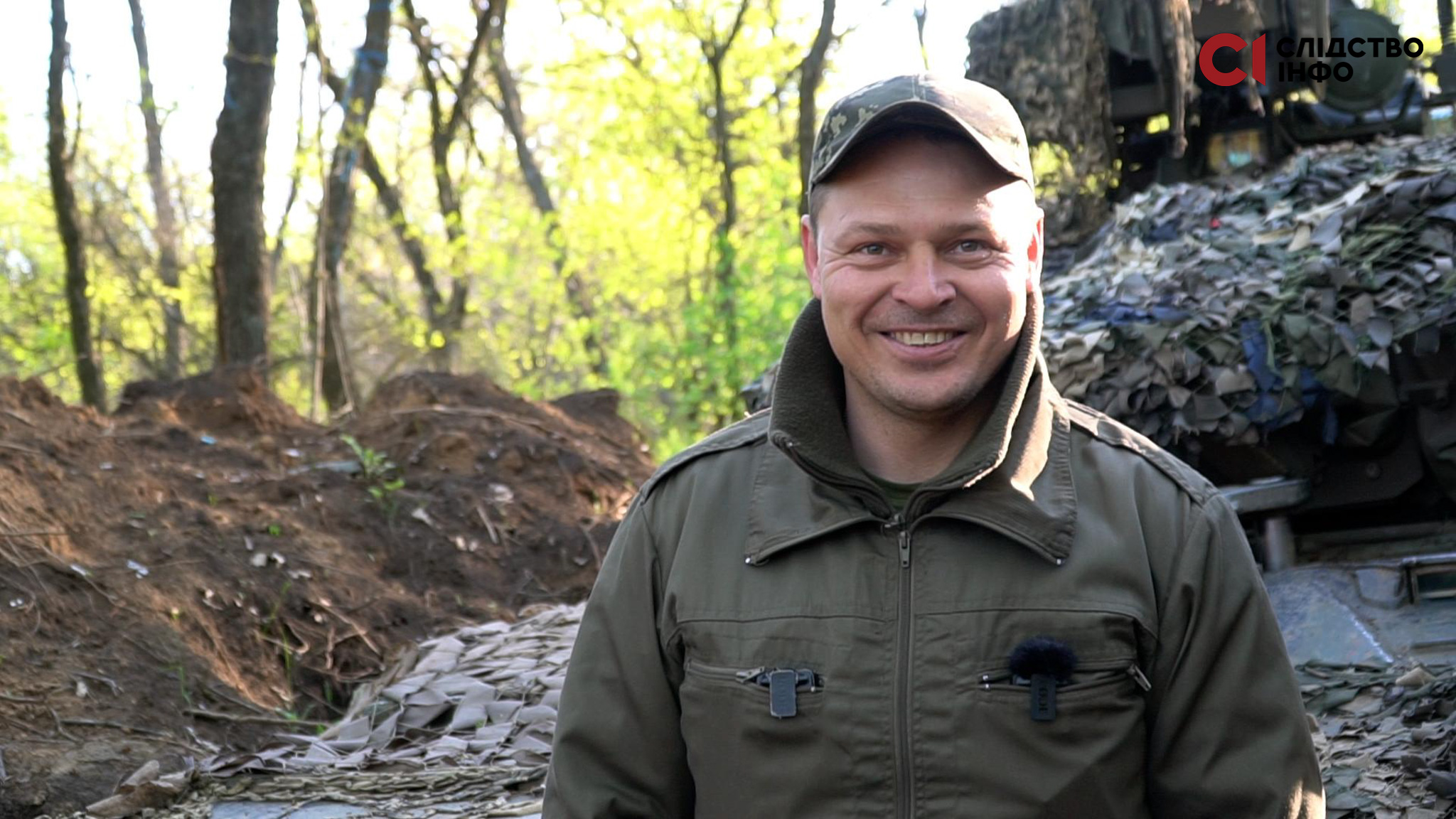 На фото чоловік у формі, стоїть у лісі біля окопів. Усміхається