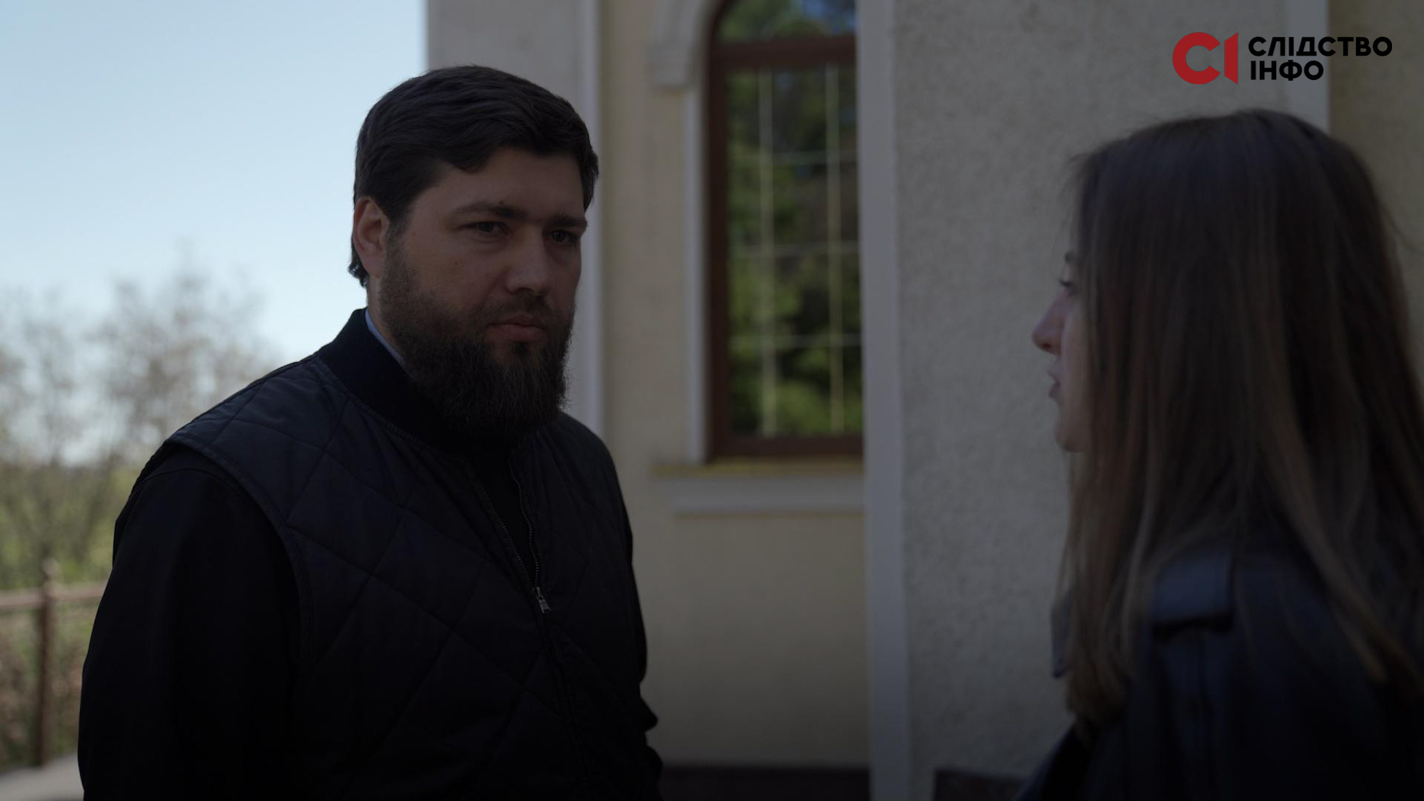На фото священник і дівчина, яка стоїть боком. Позад них будівля