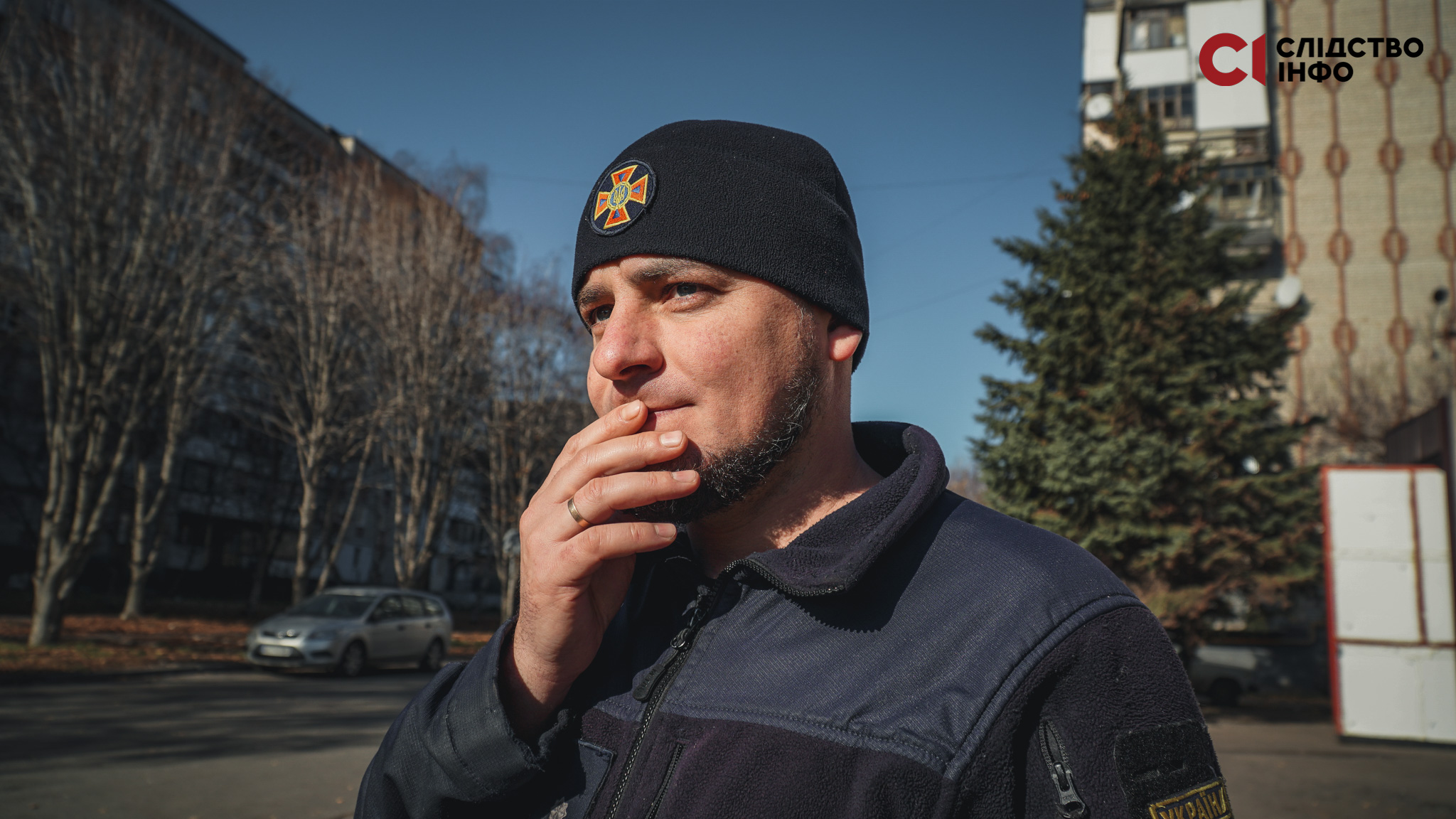 Чоловік тримає біля своїх губ руку, на фоні дерева і будинки