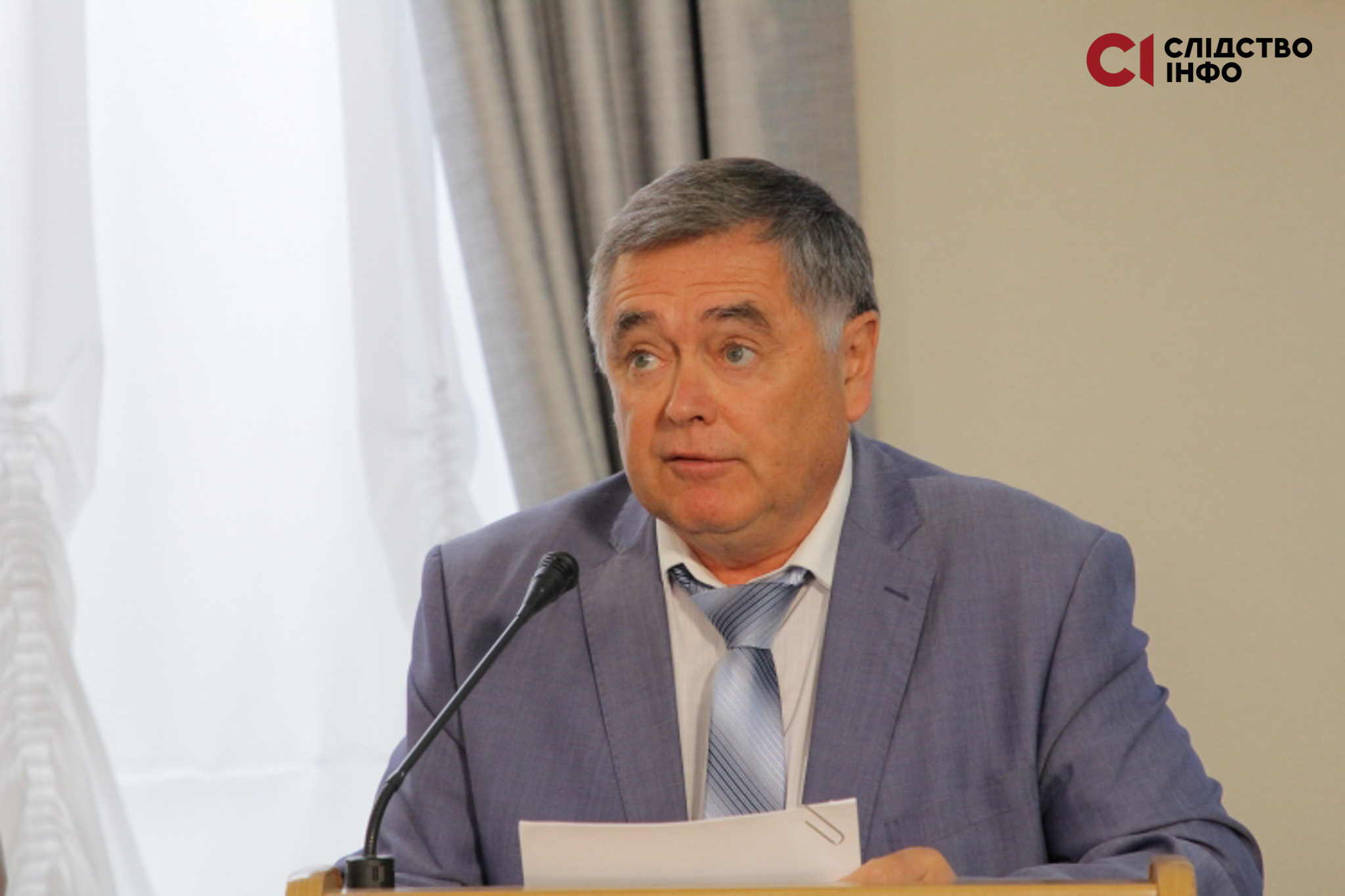 На фото чоловік у костюмі й краватці тримає у руках документ і говорить у мікрофон