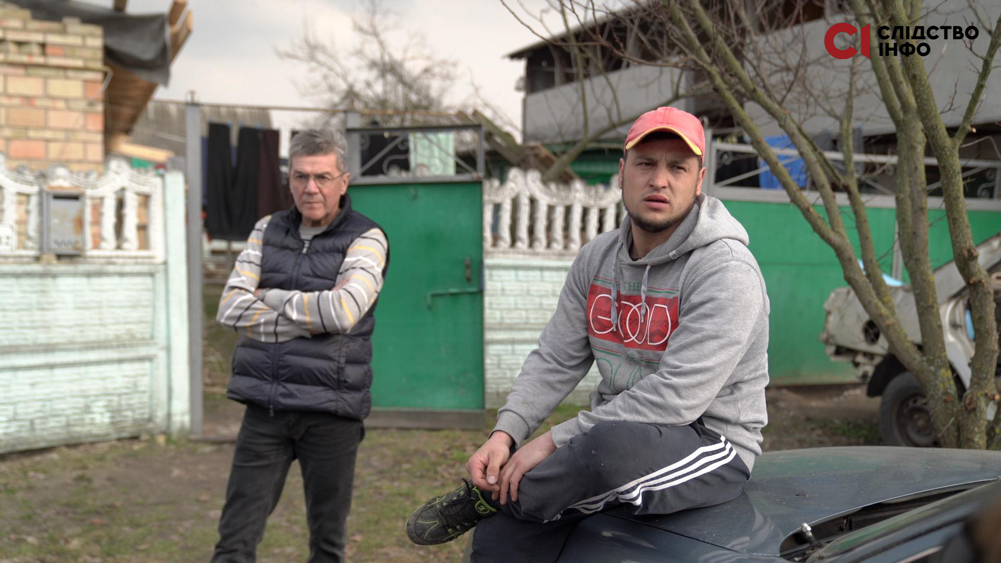 На фото два чоловіки: один стоїть на задньому фоні, схрестивши руки. Інший - сидить на капоті машини. Позад них паркан і будинки
