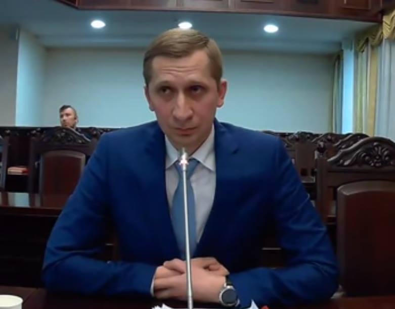 чоловік у синьома костюмі говорить у мікрофон. На фоні порожня зала з кріслами, у якій сидить лише один чоловік
