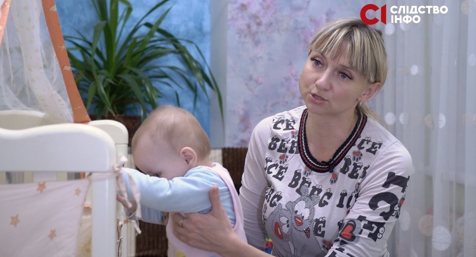 На фото жінка (справа) підтримує немовля (зліва), яке стоїть і тримається за дитяче ліжечко