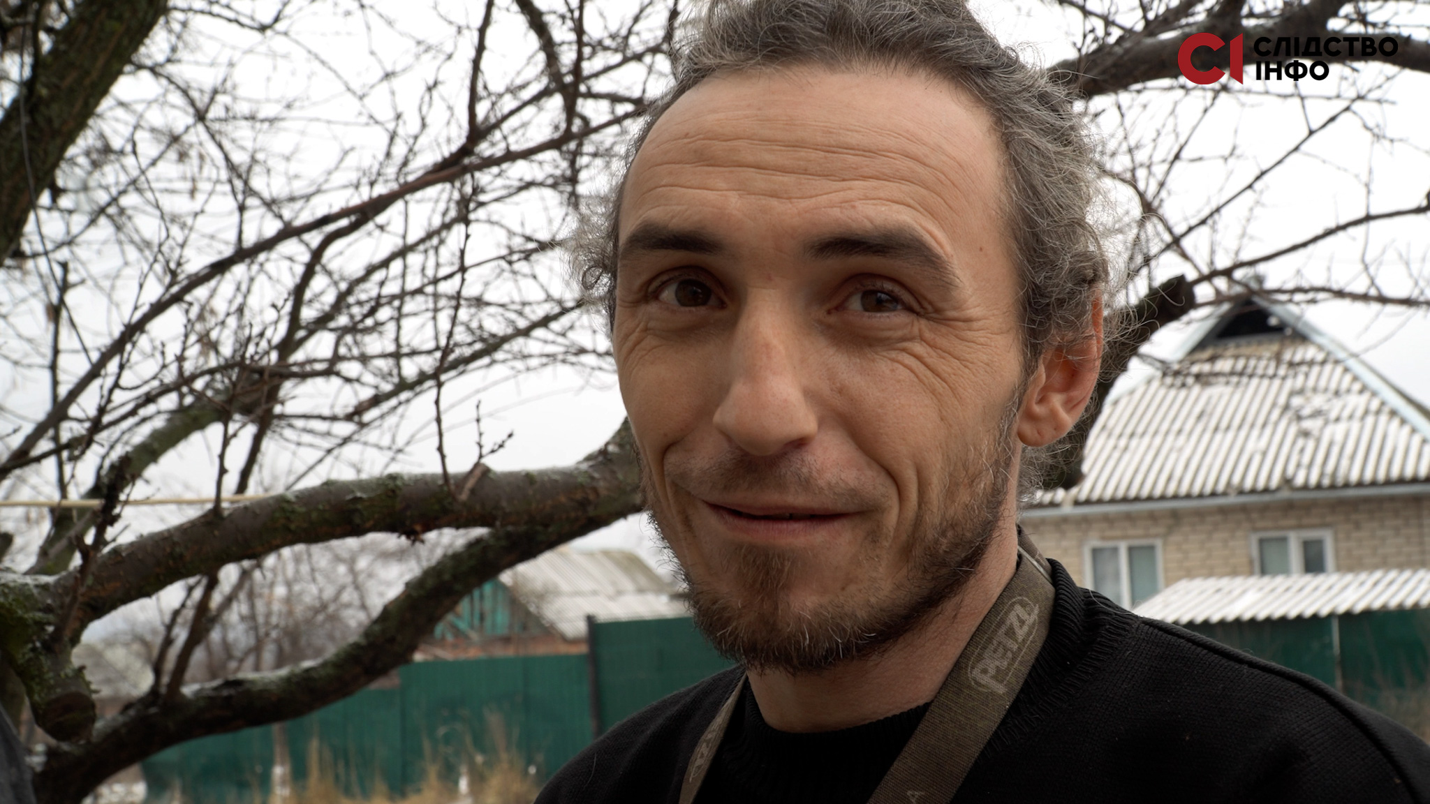 На фото крупним планом чоловік у чорному светрі, позад нього хата і гілки дерев