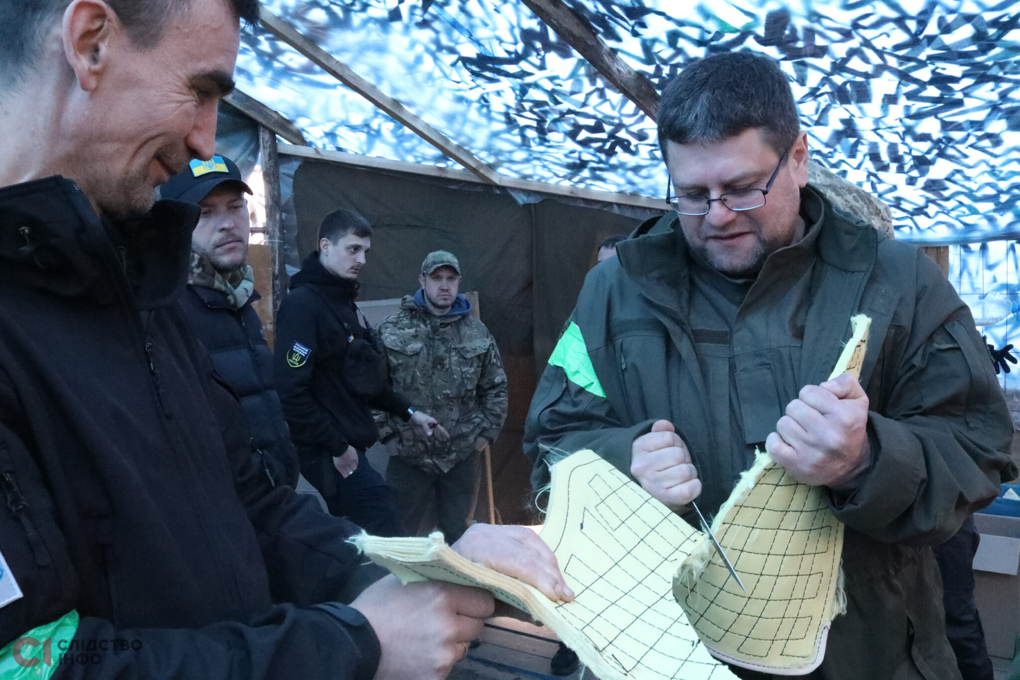 на фото чоловіки у військовій формі під стелею з маскувальної сітки. Попереду один чоловік тримає світлу цупку тканину, а інший чоловік в окулярах розрізає її ножем