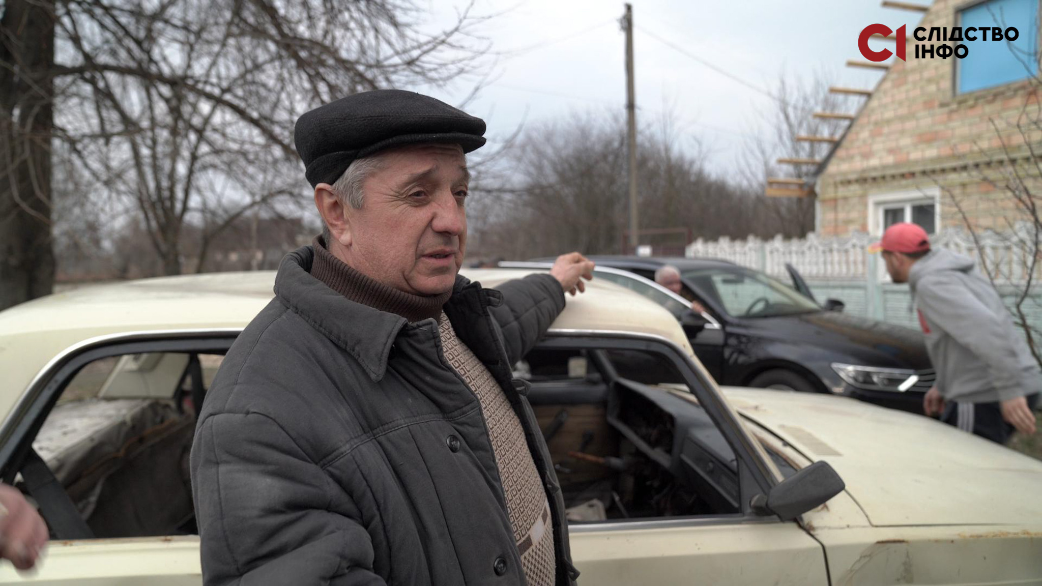 Чоловік стоїть біля авто світлого кольору й показує рукою позад себе