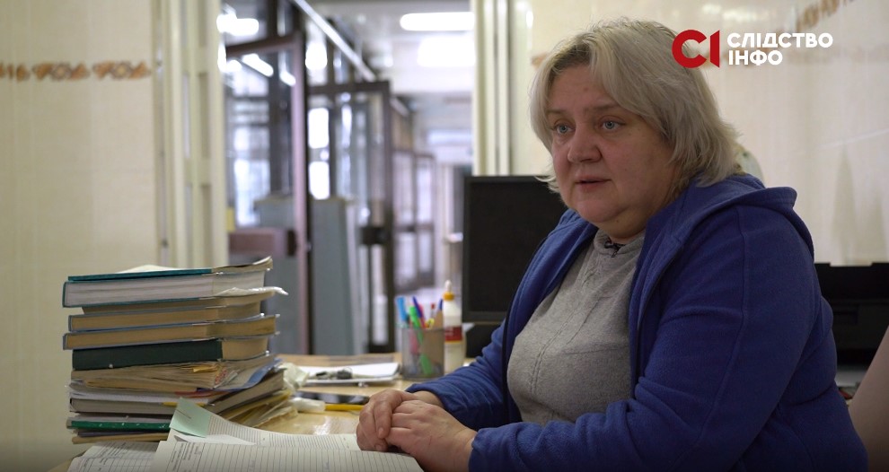 На фото білява жінка у синій кофті сидить боком за столом, на якому багато книг для записів. На задньому фоні довгий коридор лікарні з відчиненими дверима у палатах