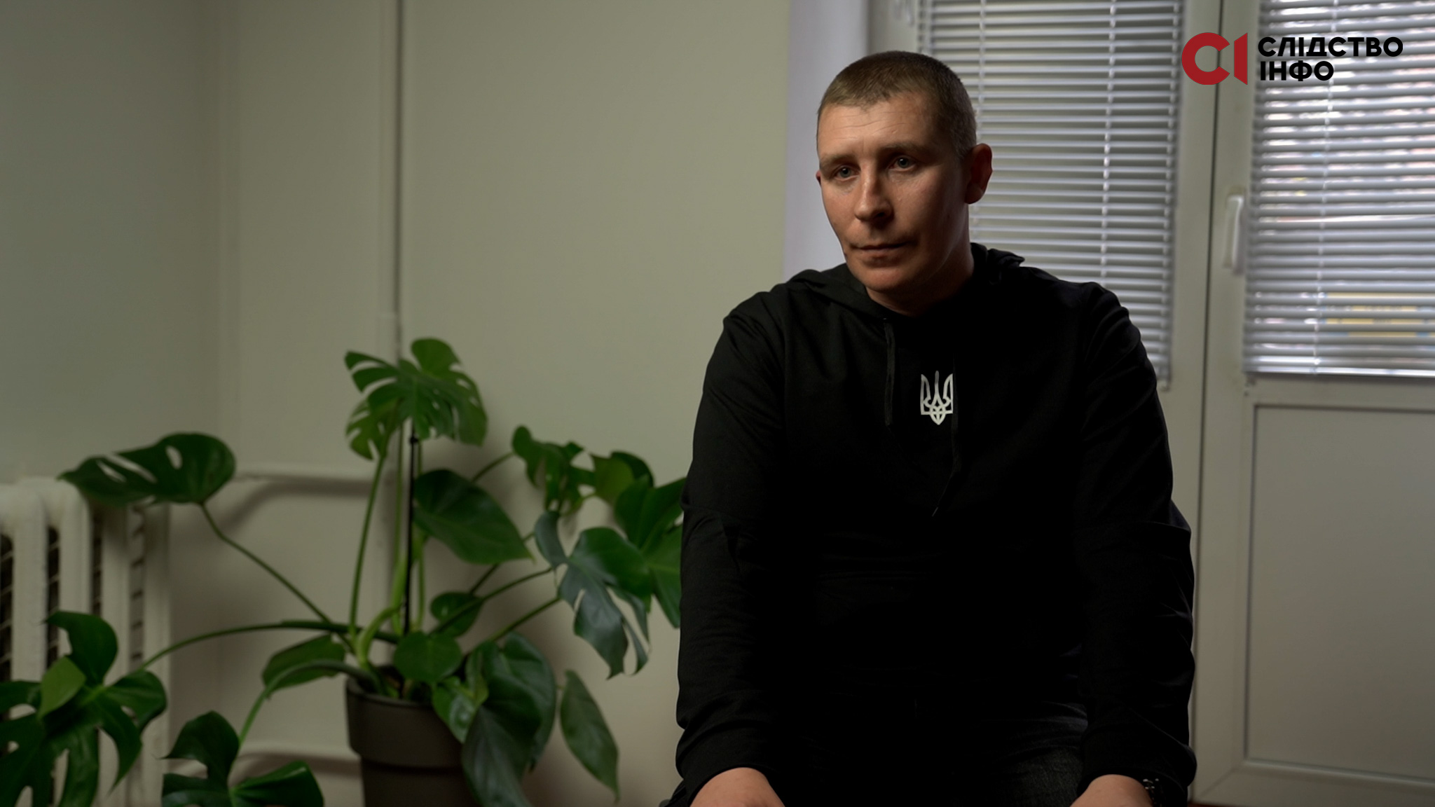 На фото чоловік у чорній толстівці з білим гербом на грудях сидить у кімнаті, позад нього велика зелена кімнатна рослина й двері на балкон