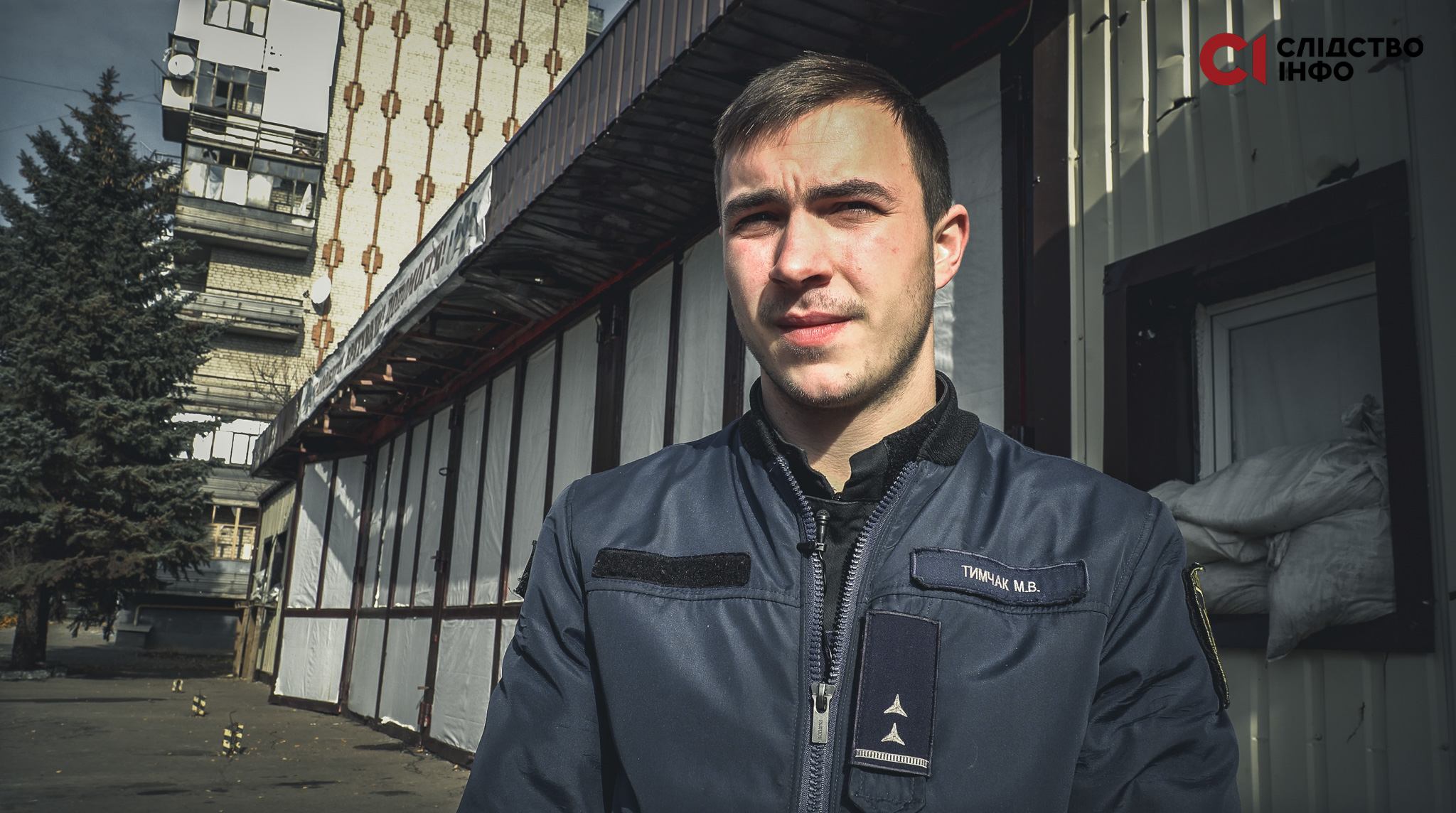Чоловік стоїть на фоні будівлі, вікно якої закладене мішками